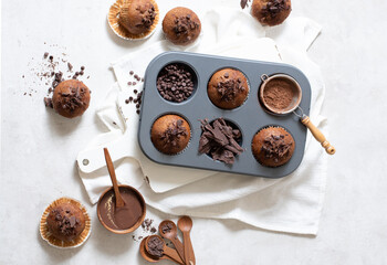 Top view of chocolate muffins flat lay in baking tray with slides of chocolate, chocolate chip, cocoa powder and chocolate sauce on white cutting board and white cloth