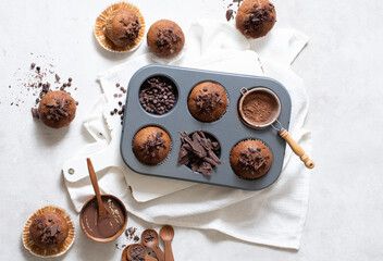 Top view of chocolate muffins flat lay in baking tray with slides of chocolate, chocolate chip, cocoa powder and chocolate sauce on white cutting board and white cloth