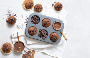 Fototapeta na wymiar Top view of chocolate muffins flat lay in baking tray with slides of chocolate, chocolate chip, cocoa powder and chocolate sauce on white cutting board and white cloth
