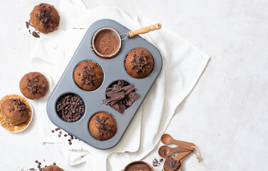 Top view of chocolate muffins flat lay in baking tray with slides of chocolate, chocolate chip, cocoa powder and chocolate sauce on white cutting board and white cloth with copy space