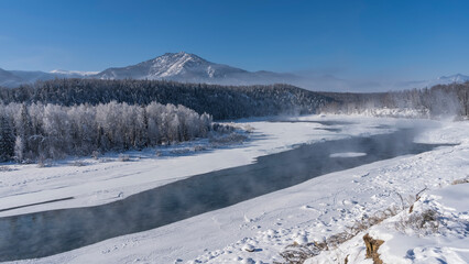 An ice-free river flows through a snow-covered valley. Steam over turquoise water. The trees are covered with frost. A picturesque mountain range against the blue sky. Altai. Katun