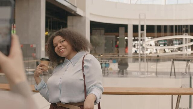 Slowmo of young curvy Black woman smiling while standing at modern shopping mall with takeaway coffee and many paper shopping bags in hands and her female friend taking pictures of her on smartphone