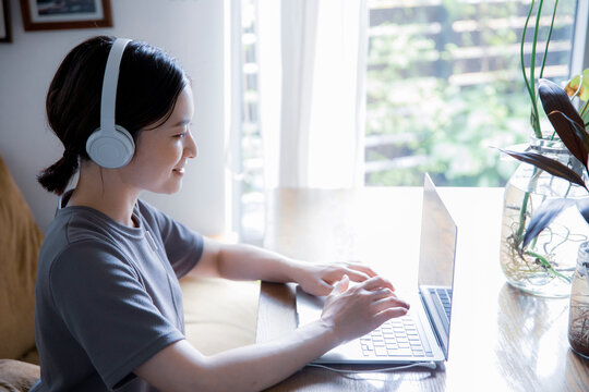 明るい植物のある窓辺の部屋でノートパソコンでオンライン授業,リモートワークをする日本人女性