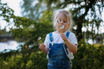 Pretty little girl blowing off dandelion seeds on sunset in summer park. Image with selective focus