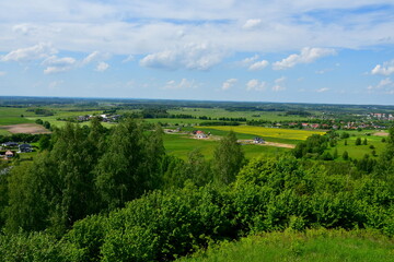 A view from the top of a tall hill or mountain with vast fields, meadows, forests, moors, and other types of flora visible, with a lake, some urban settlements, and other buildings in the distance