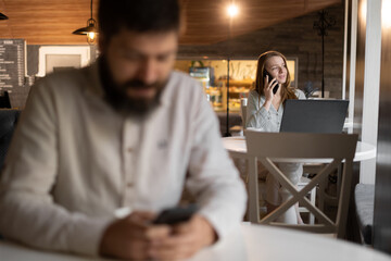 The girl looks at the profile page of a handsome man, browses a dating site, sitting in a cafe with a laptop makes a call on a phone, selective focus man in the foreground