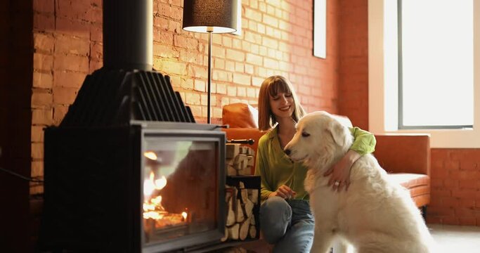 Young woman feeds her white adorable dog, spending time together in cozy living room with fireplace at home. Concept of friendship with pets