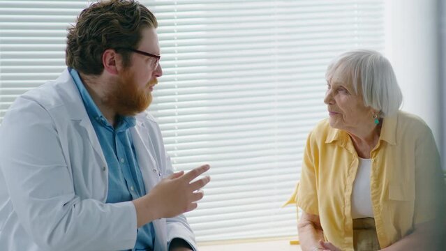 Elderly Caucasian woman listening to young male doctor while getting medical consultation in clinic