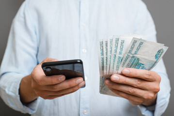 Business, finance and technology concept. Close-up of man holding thousands of rubles and using smartphone while standing indoors on gray background. Selective focus on money and mobile phone