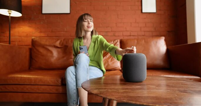 Young woman using phone and controlling home devices with a voice command while sitting relaxed on leather couch. Concept of smart home and leisure time with gadgets