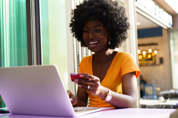 Happy young african woman holding credit card while shopping via laptop sitting in outdoor cafe