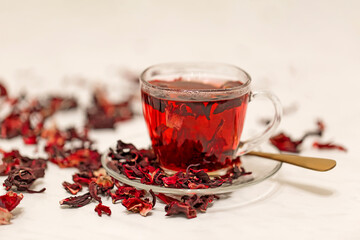 Red hot hibiscus karkade tea in a glass mug on a white table among dried flowers of the Sudanese rose.