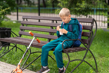 Fototapeta premium a boy in a blue sweatshirt sits on a bench in the park and eats ice cream from a cup