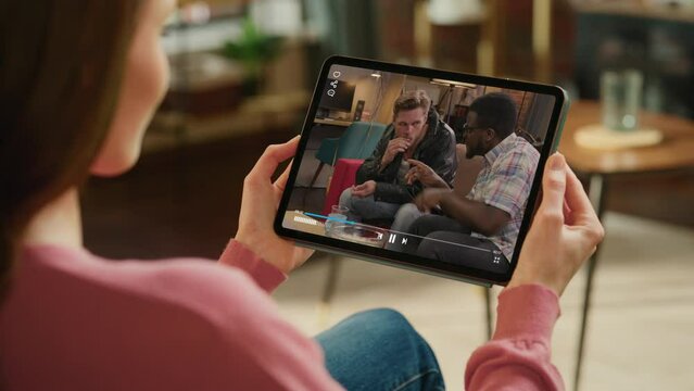 Over Shoulder Woman Using Digital Tablet Computer Watching Hilarious Popular Sitcom on TV. Girl Have Fun on a Couch Watching Hit Comedy TV Series about Funny Group of Friends On Streaming Service