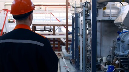 Work at the factory, a worker in a helmet and overalls walks past the equipment. The worker makes a tour of the equipment to check the condition of the equipment. View from the back.