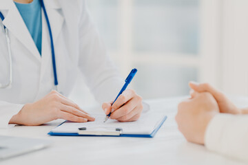 Unknown female doctor writes down prescription on papers in clipboard, consults patient about curing disease, pose at desktop against blurred white background fills up medical form. Symptoms diagnosis