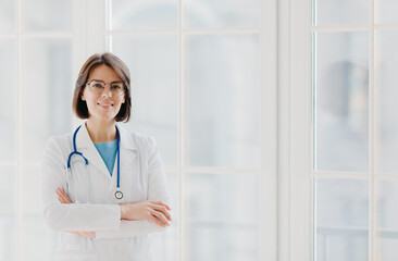 Self confident professional doctor stands with arms crossed, wears white medical gown with stethoscope, thinks about work positively, poses against big window. Healthcare and occupation concept