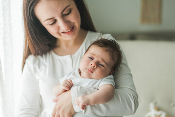 Close portrait of a little cute dark-haired newborn girl in a white suit in the arms of a young mother. Motherhood. Parent. Healthy sleep.