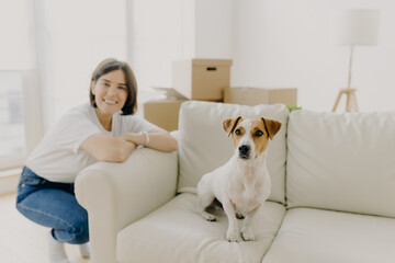Happy woman plays with favourite pet, poses near sofa in new apartment, celebrate Moving Day,...