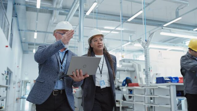 Portrait of a Two Diverse Heavy Industry Engineers in Hard Hats Walking with Laptop Computer and Talking in a Factory. Slow Motion Footage of Two Manufacturing Employees at Work.