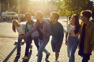 Exploring city together. Goup of cheerful friends walk streets of new city together during weekend vacation. Active millennial multiracial casual women and men laugh and talk while walking down street - obrazy, fototapety, plakaty