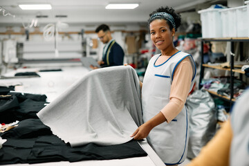 Obraz premium Happy African American seamstress works with fabric at textile workshop and looking at camera.