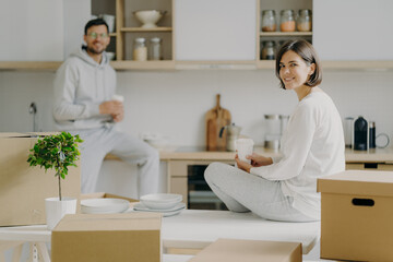 Obraz premium Horizontal shot of happy young woman and man sit on kitchen desk, drink coffee, have break after unpacking boxes, have pleasant talk, surrounded with plates, indoor plant in pot. Moving concept