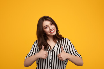 Cheerful young woman showing thumbs up in yellow studio