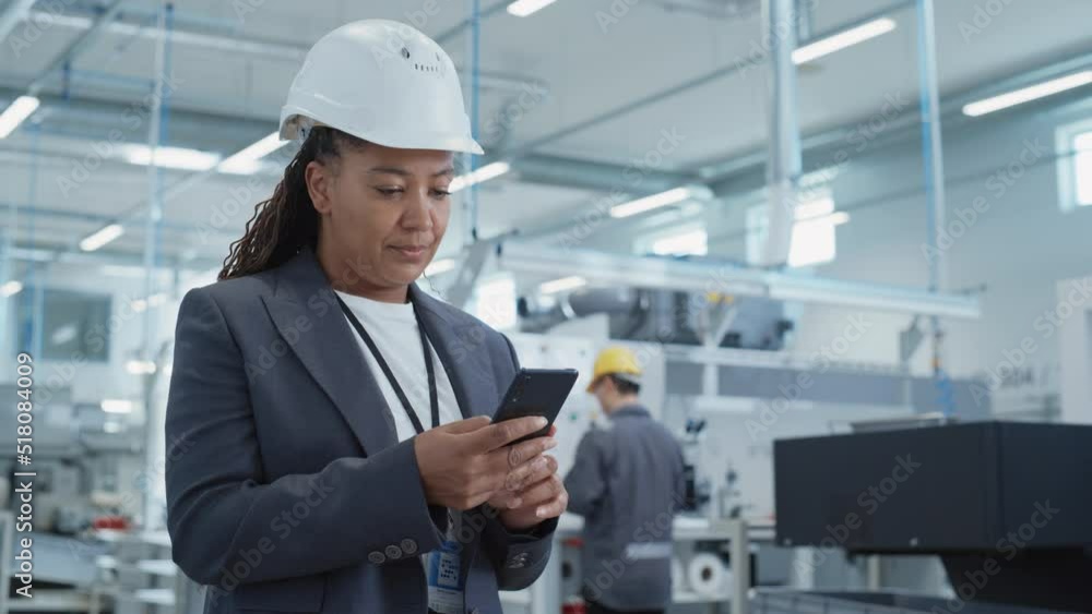 Fototapete Portrait of a Black Female Engineer in Hard Hat Standing and Using a Smartphone at Electronics Manufacturing Factory. Technician is Writing a Message and Checking Her Schedule.