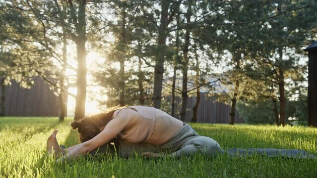 Woman doing yoga in yard. Young brunette stretching in Trianga Mukha Eka Pada Paschimottanasana and Ardha Matsyendrasana poses during yoga session on green grass in summer morning in yard
