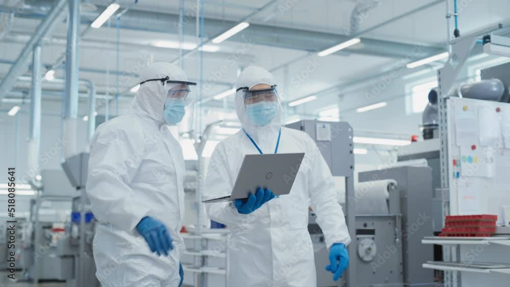 Poster two scientists standing in a heavy industry factory in sterile coveralls and face masks, using lapto