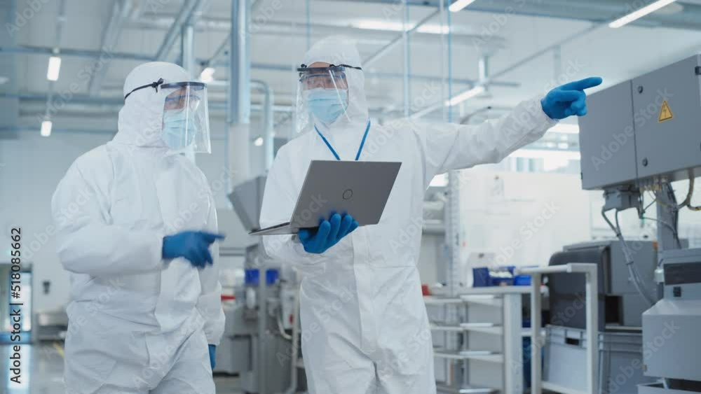 Poster two scientists walking in a heavy industry factory in sterile coveralls and face masks, using laptop