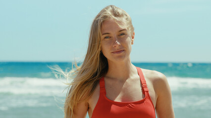 Portrait of beautiful young woman in red swimsuit stand on empty sunny beach, look into camera with confidence. Baywatch or coast guard tower in background. Summertime concept. Warm breeze in hair