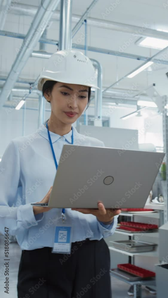 Poster Vertical Portrait of an Asian Female Engineer in Hard Hat Walking and Using Laptop Computer at Electronic Manufacturing Factory. Technician Working on Daily Tasks and Research and Development Data.