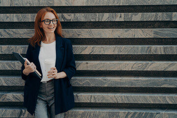 Woman dressed formally drinks takeaway coffee holds digital tablet and notepad