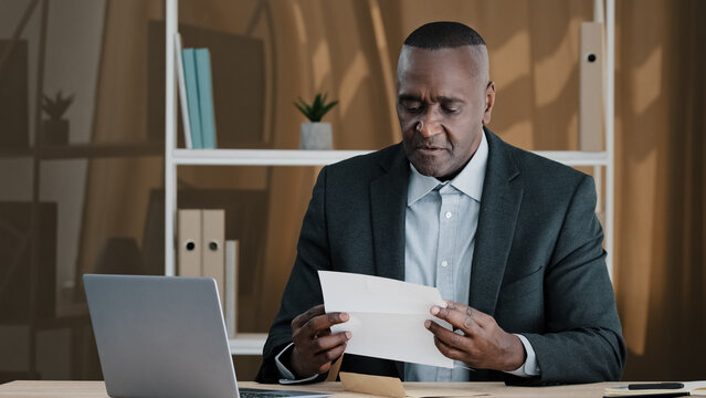 African old man senior mature male in office opening paper envelope reading letter with good news. Happy elder retired businessman received investment notification banking loan approval salary bonus
