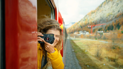 Inspiring female traveler and travel blogger look out of window of tourist train in cinematic beautiful mountains. Young beautiful woman hang out of window of red train. Excited world explorer