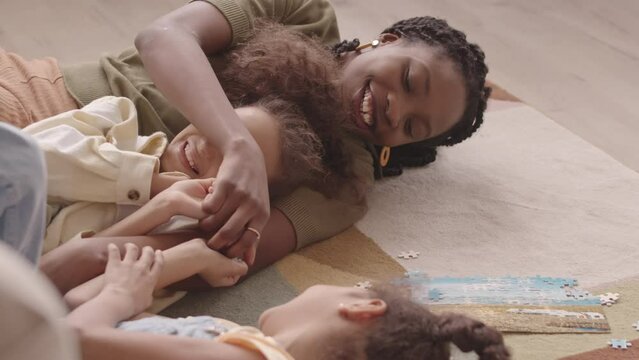 Slowmo of cheerful African American woman and her two pretty little daughters lying on floor having fun while solving puzzles together at home