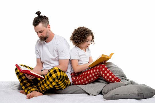 Family in plaid sleepwear sit on bed and read paper books on white studio background. Dad and curly short-haired daughter in pajama. Daddy and female child girl spend time together for regtime bedtime