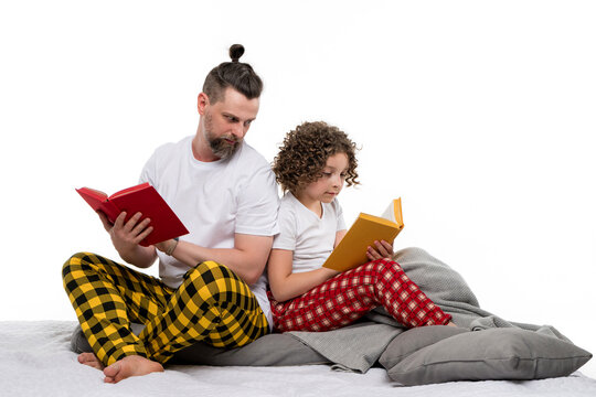 Family in plaid sleepwear sit on bed and read paper books on white studio background. Daddy and curly short-haired daughter in pajama spend time together for regtime bedtime. Dad looks at child book