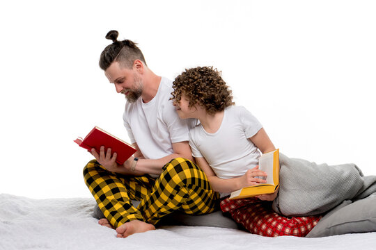 Family in plaid sleepwear sit on bed and read books on white studio background. Dad and curly short-haired daughter in pajama spend time together for regtime bedtime. Child looks at father book