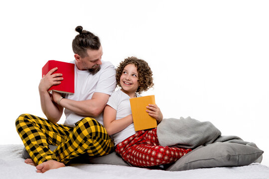 Family in plaid sleepwear sit on bed and read books on white studio background. Dad and curly short-haired daughter in pajama having fun together. Leisure concept for regtime bedtime.