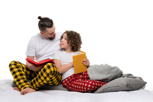 Funny Family in plaid pajamas sit on bed and read books on white studio background. Single Dad and curly short-haired daughter in sleepwear pajama show tongue to each other. Leisure regtime bedtime