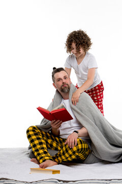 Happy family in plaid pajamas on white studio background. Caring curly short-haired daughter in sleepwear covering dad with blanket. Daddy reading book. Leisure regtime bedtime. 