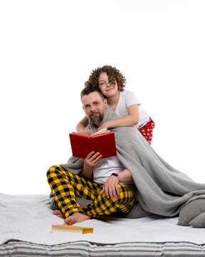 Happy family in plaid pajamas on white studio background. Caring curly short-haired daughter in sleepwear hugging dad with blanket. Daddy reading book. Leisure regtime bedtime. 
