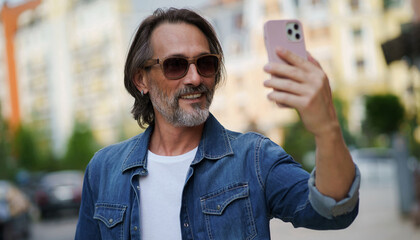 Handsome stylish sexy middle aged grey bearded man smiling on camera old town wearing casual. Happy senior man on street of european city making selfie using mobile phone. Travel concept