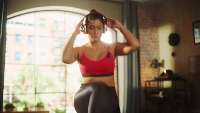 Beautiful Athletic Plus Size Body Positive Sports Woman Uses Smartphone Health Fitness Tracking App, Does Aerobic Exercises, Squatting, Knees to Elbows at Home. Female Training Wearing Headphones