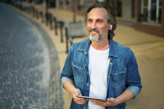 Traveling handsome middle aged man using digital tablet having a call while standing on urban city streets. Mature man talking or listening music use wireless earphones while travel old town streets - Powered by Adobe