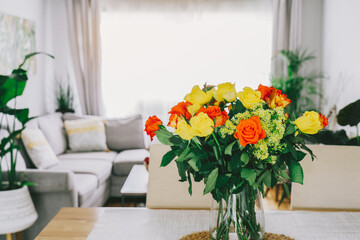 Kitchen counter table with focus on vase with yellow and orange roses on it with blurred background of modern cozy living room with couch and green plants. Open space home interior design. Copy space.