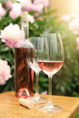 Glasses and bottle with rose wine on wooden table near beautiful peonies